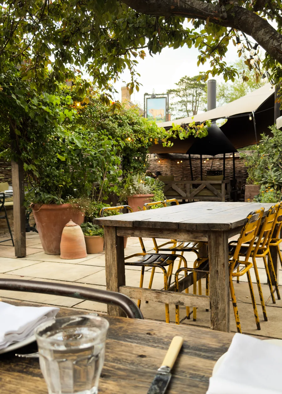 The Bull's garden terrace in Charlbury, with weathered timber tables, yellow chairs and a wood-fired oven under fruit trees and festoon lights