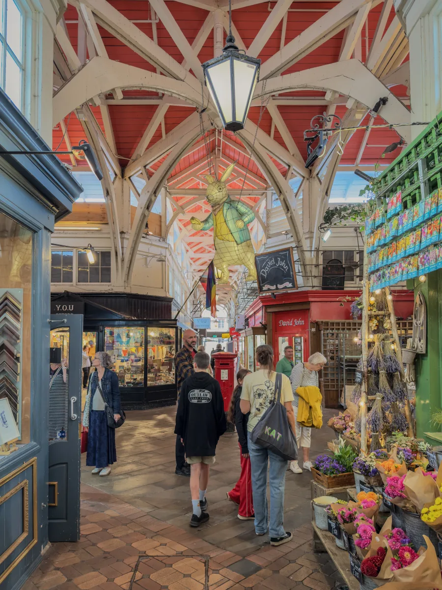 A flower stall banked with lavender and bouquets along an avenue of Oxford Covered Market, the hanging White Rabbit sculpture from Alice in Wonderland above