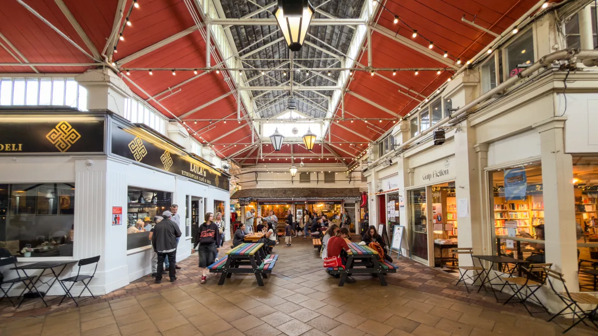 The main hall of Oxford Covered Market under its red-timbered roof, with delis, a bookshop and striped picnic benches