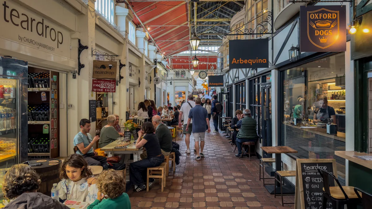 A busy café-lined avenue inside Oxford Covered Market, with diners at tables under the glass and timber roof