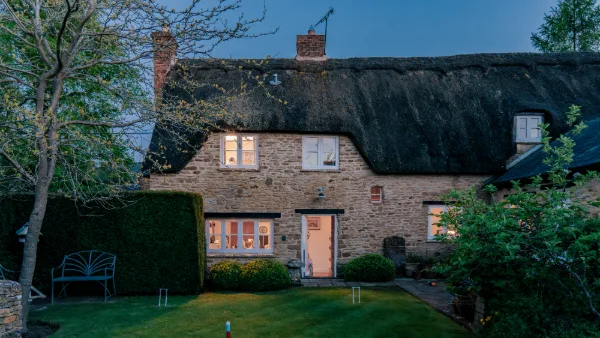 Well Cottage from the garden at twilight, thatched roof silhouetted and windows glowing warm