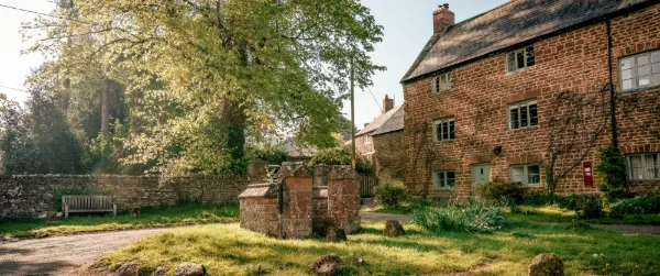 Well Cottage from the lane in soft morning light, ironstone walls and trees overhead