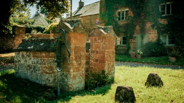 The old stone well and iron pump on the village green, the ivy-clad cottage behind