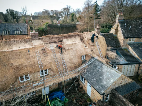 Two thatchers stripping old straw from the roof, the cottages of Ledwell behind