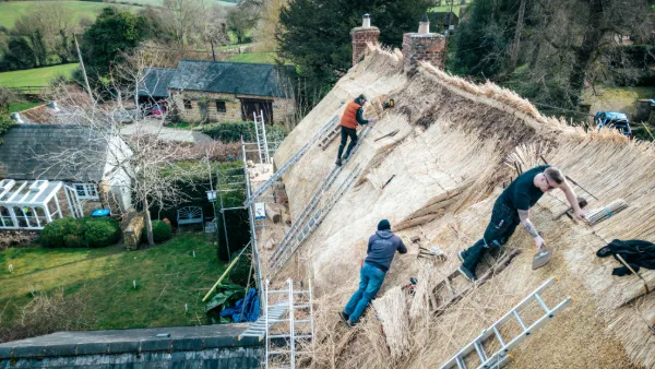 Three thatchers working the roof from ladders, the walled garden and greenhouse below