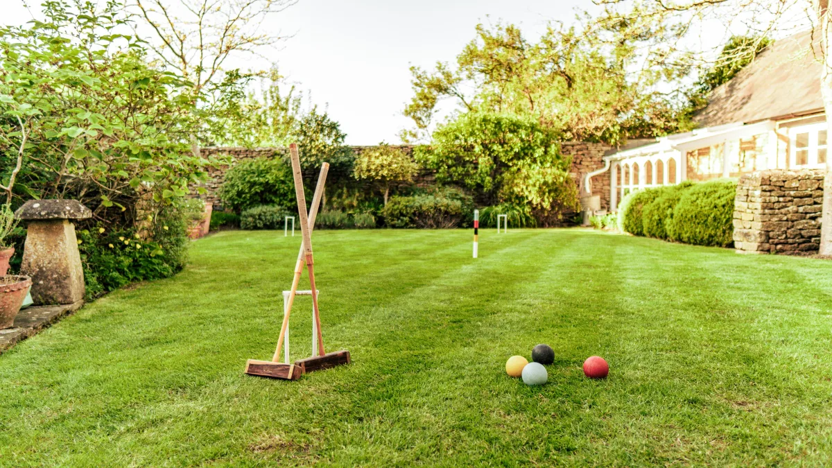 Black, green, red and yellow croquet balls in the grass beside a hoop, a mallet leaning on its stand and the conservatory catching the evening light beyond