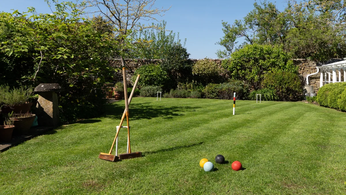 A croquet mallet resting on its stand on the sunny lawn, hoops and three coloured balls laid out ready to play, the dry-stone wall behind