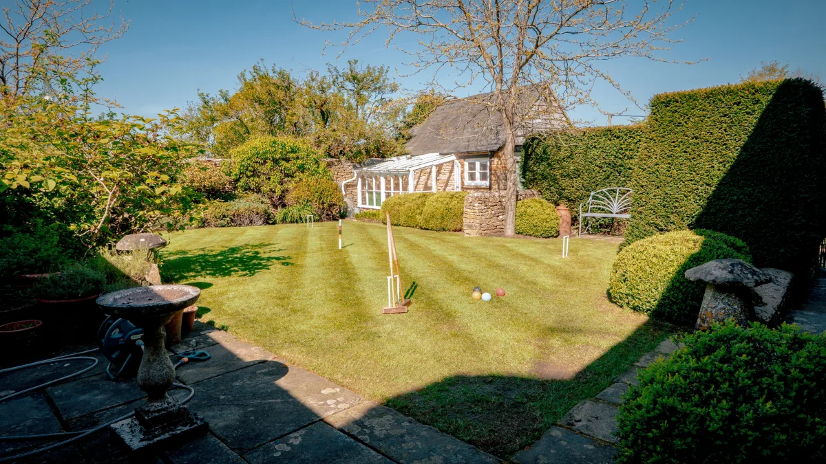 The garden seen from the stone patio at Well Cottage, the sunlit striped lawn sweeping up to the thatched cottage and conservatory with a tall clipped hedge alongside