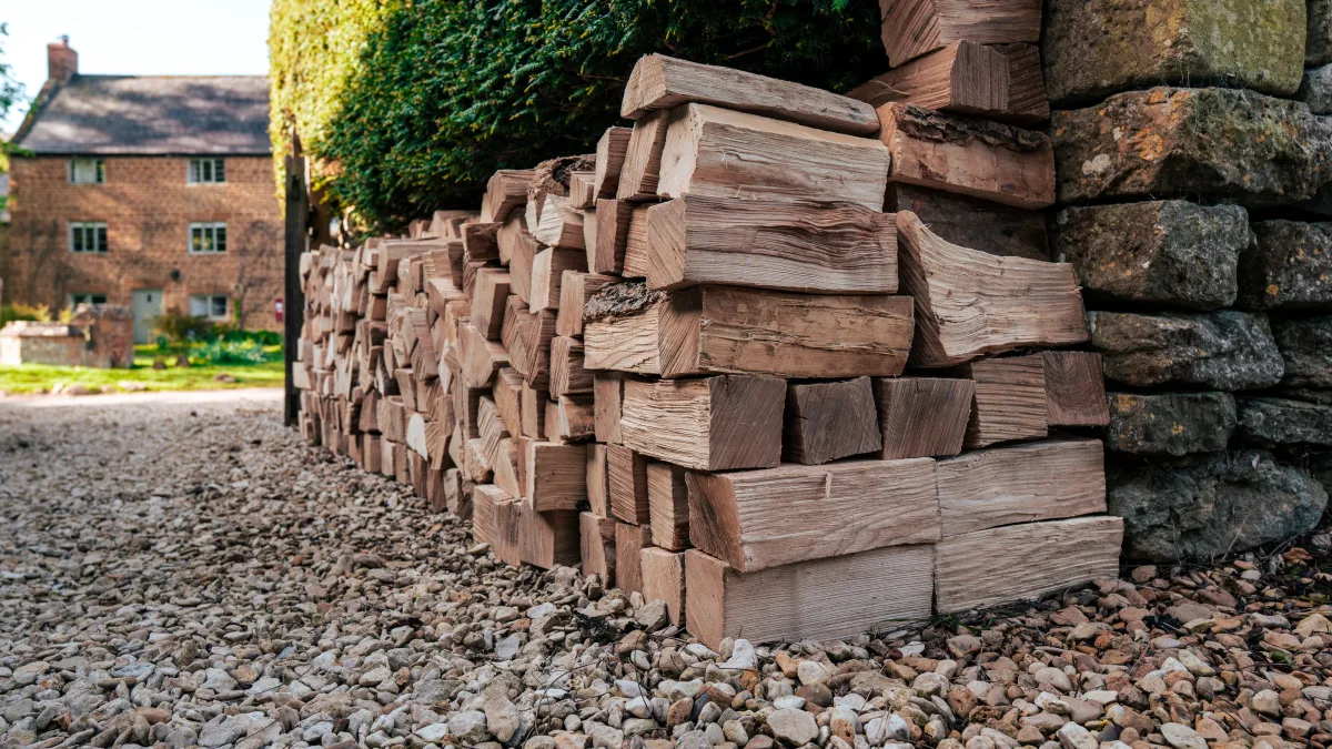 A close-up of the stacked log store at Well Cottage on its bed of gravel, the honey-stone cottage and a clipped hedge behind