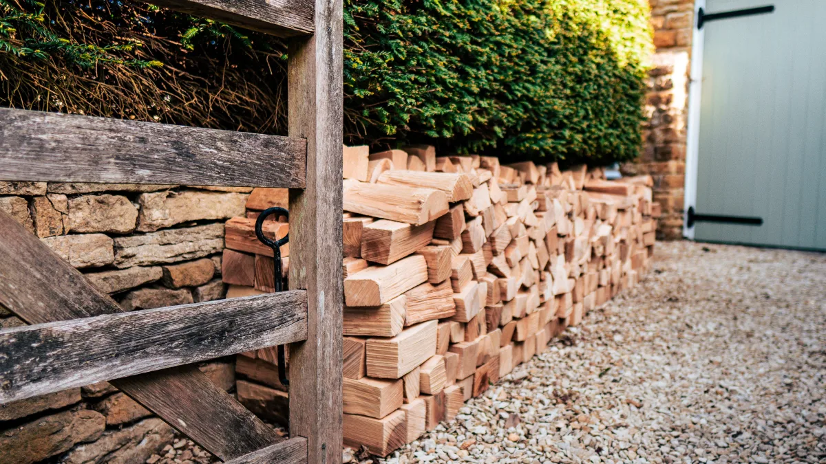 A neat run of split logs stacked along the gravel path at Well Cottage, beside a weathered timber gate and clipped hedge