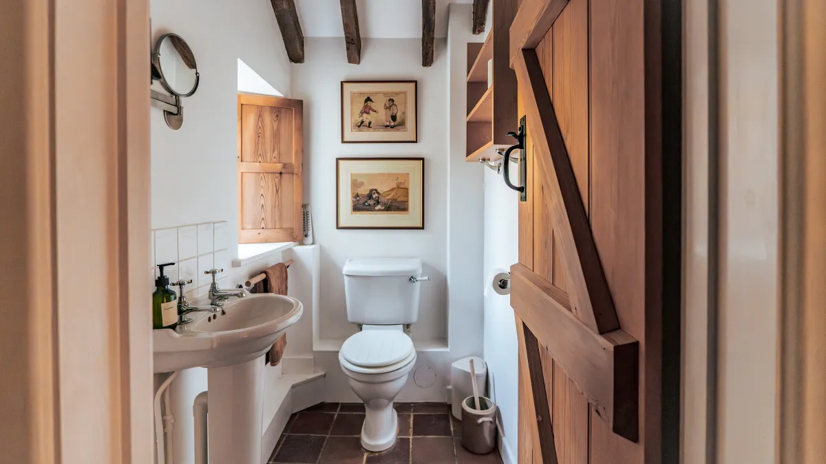 The downstairs bathroom at Well Cottage — exposed ceiling beams, a pedestal basin, framed antique prints and a pine plank door on a wrought-iron latch, over a terracotta-tiled floor