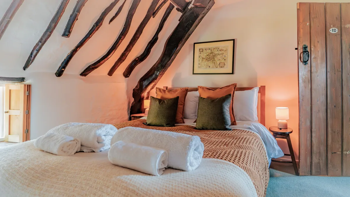 The king bed seen from the foot in warm lamplight, towels rolled at the end, olive and ochre cushions against the headboard and the numbered plank door to one side