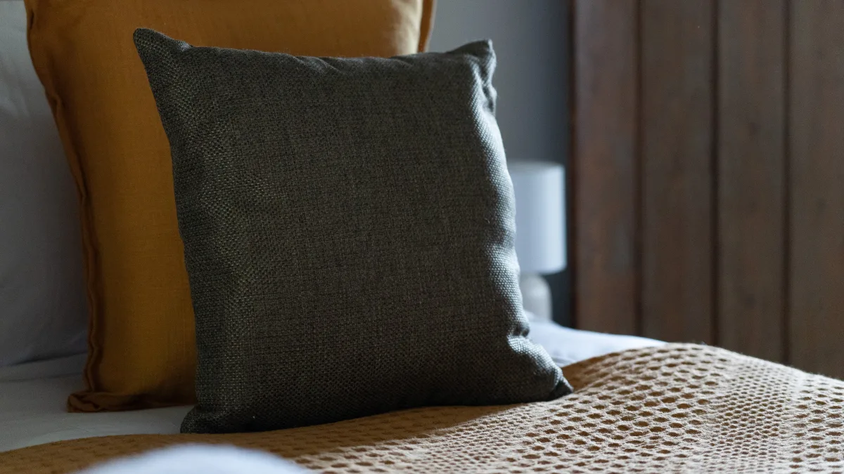 A charcoal-olive linen cushion propped against an ochre cushion on the waffle bedspread, the bedside lamp soft beyond