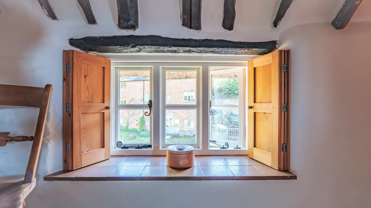 Pine shutters folded back from a deep three-light cottage window in the King Room, looking out over the thatched stone cottages of Ledwell