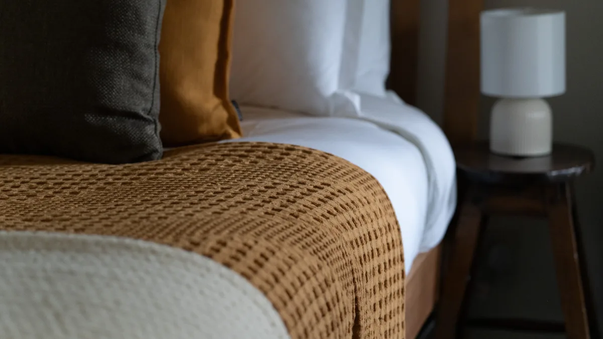 The ochre waffle throw draped over crisp white linen at the foot of the king bed, a turned-wood stool with a lamp just beyond