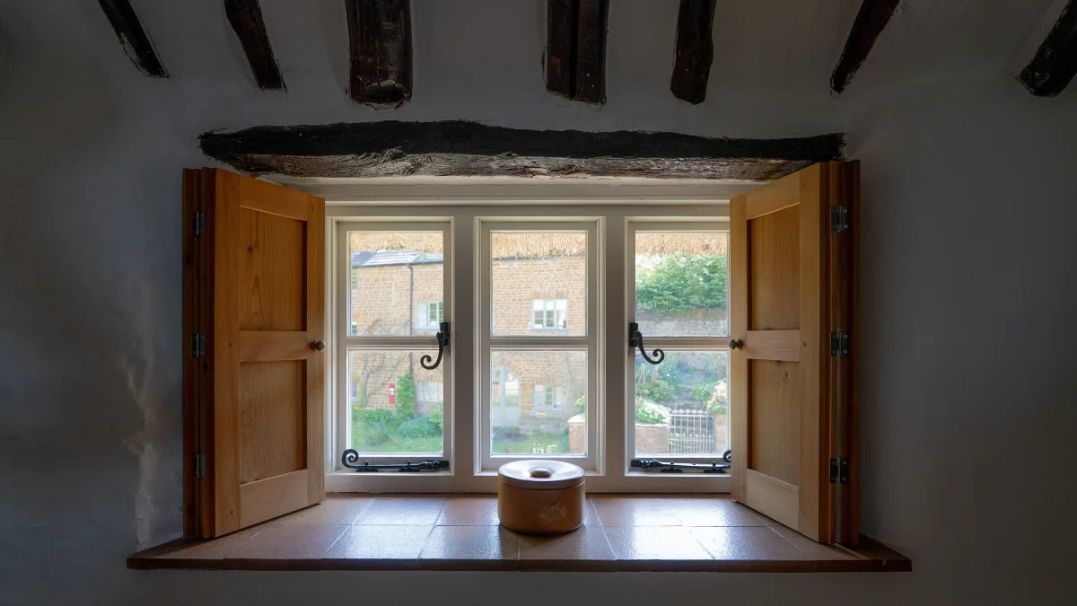 The deep recessed window of the King Room with its pine shutters open under a great oak beam, looking out to thatch and brick across the lane