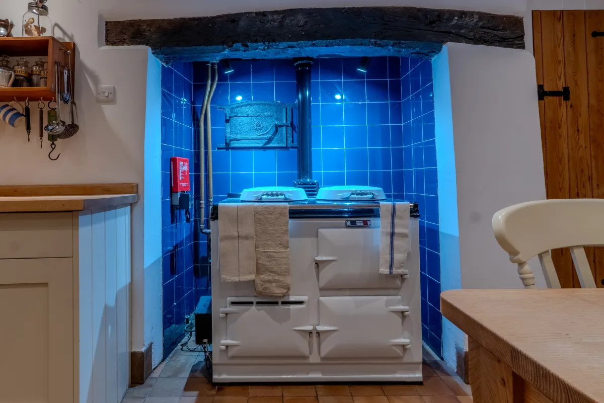 The top of the Aga seen close, its two enamelled hotplate lids and chrome rail against the deep-blue glazed tiles, the old cast-iron flue door and copper pipework above