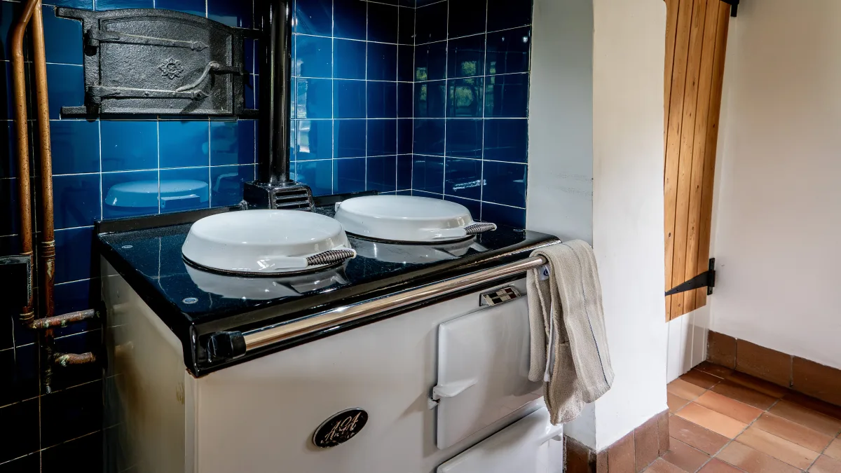 The Aga's black granite top with its two domed hotplate lids and chrome rail, a striped linen cloth hung over the rail and copper pipes running up the blue-tiled wall