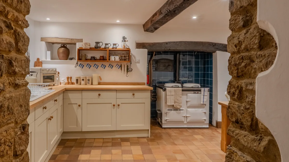 The kitchen seen through the stone arch in the evening, the cream cabinetry and open shelves to the left and the Aga lit in its blue recess beyond, beams overhead