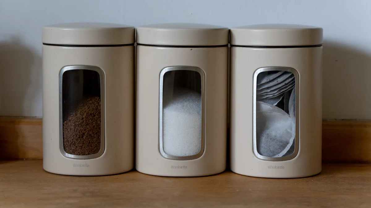 Three cream canisters with little viewing windows showing coffee, sugar and tea bags, lined up on the maple worktop