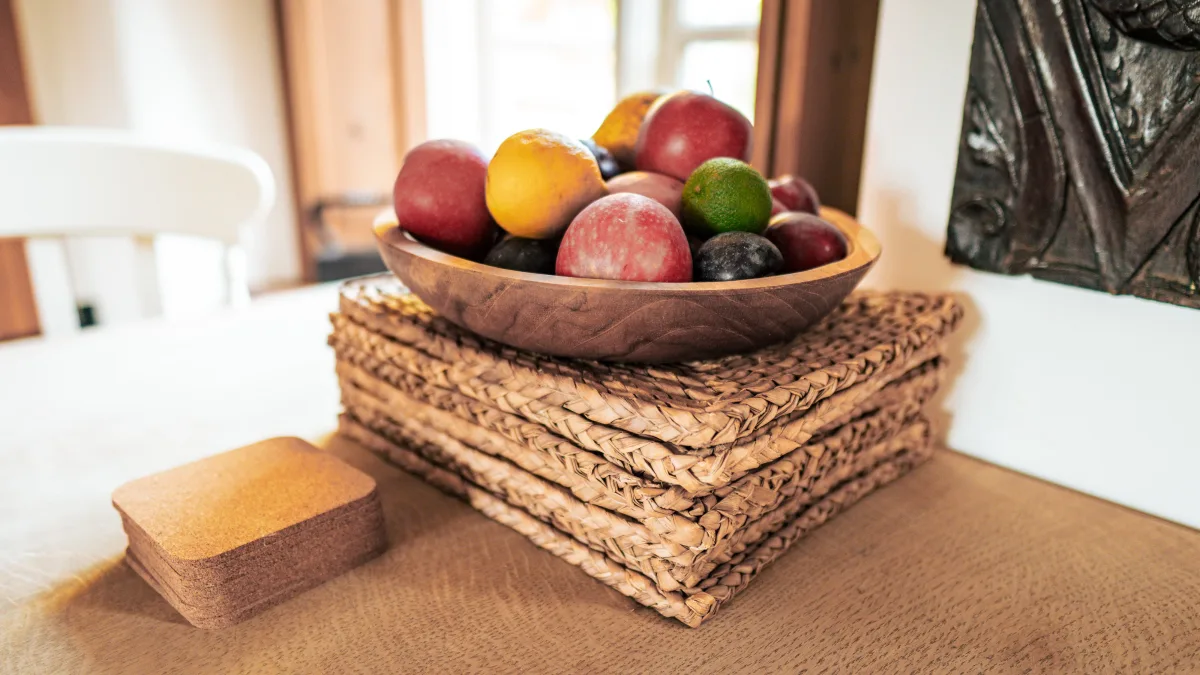 A turned wooden bowl of apples, plums and a lime sitting on a stack of woven placemats on the oak table, cork coasters beside it and a carved panel on the wall behind
