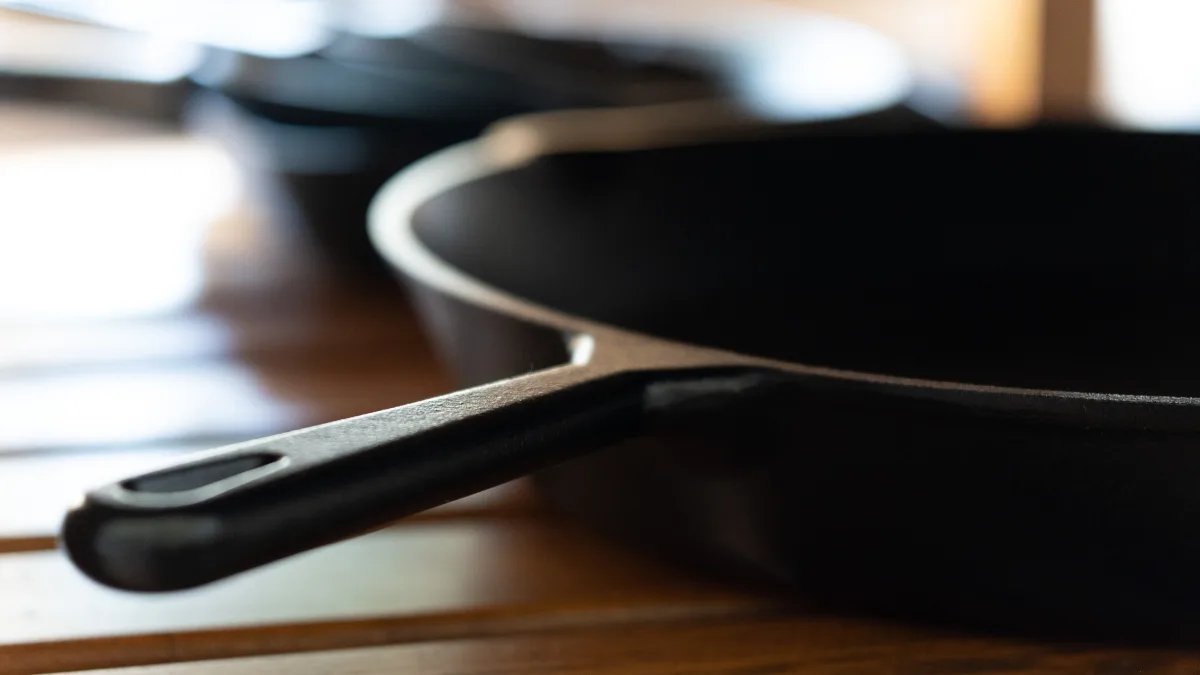 A heavy cast-iron skillet resting on a wooden surface, its long handle in sharp focus and a second pan soft behind