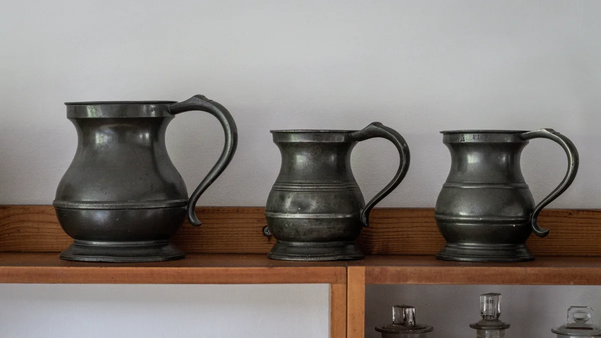 Three antique pewter tankards in graded sizes standing on the wooden shelf, with little glass apothecary jars visible on the shelf below