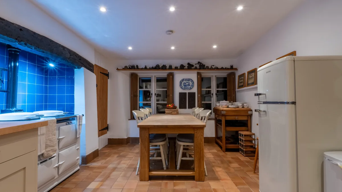 The full length of the kitchen in the evening, the laid oak table and dark carver in the foreground, the Aga glowing blue to the left and the cream fridge and dresser to the right under the downlights