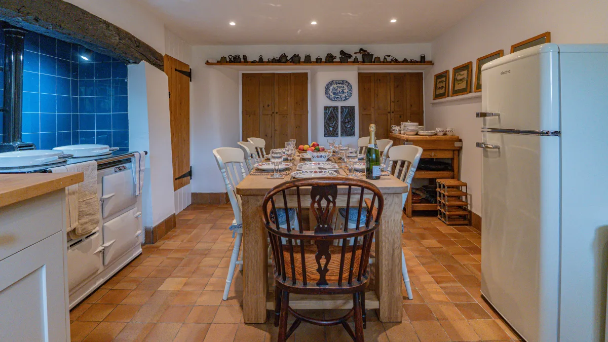 The kitchen from the table end in the evening, the laid oak table and dark carver in front, the blue Aga recess to the left and the cream retro fridge and pine cupboards to the right