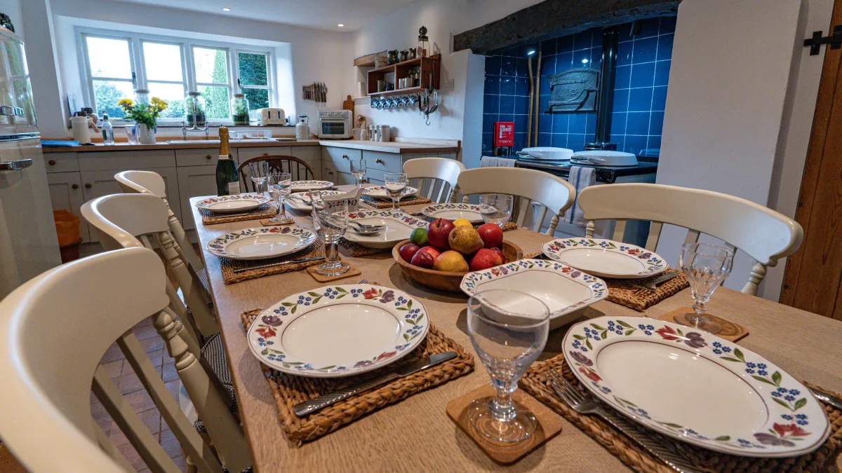 Place settings running down the length of the oak table — floral plates on woven mats, cut glasses on cork coasters and a bowl of fruit — with the garden window and the kitchen beyond