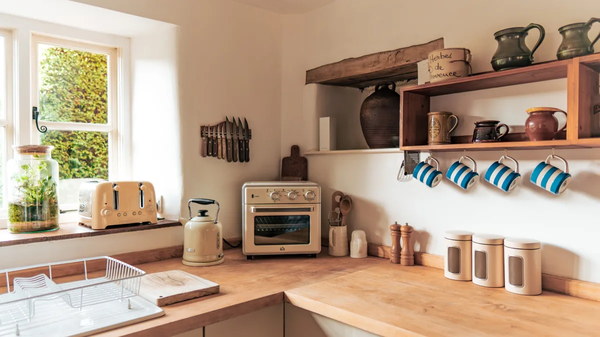 The corner of maple worktop with the cream toaster, kettle and stainless worktop oven below a wall of knives, a row of hanging striped mugs and the open shelf of jugs