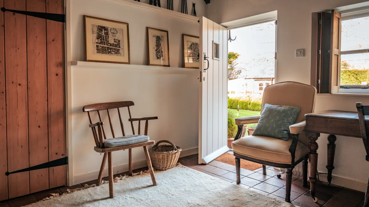 A corner of the library with the plank door open to the garden, an old country pine chair, framed Ledwell prints on the picture shelf and the cream armchair by the window