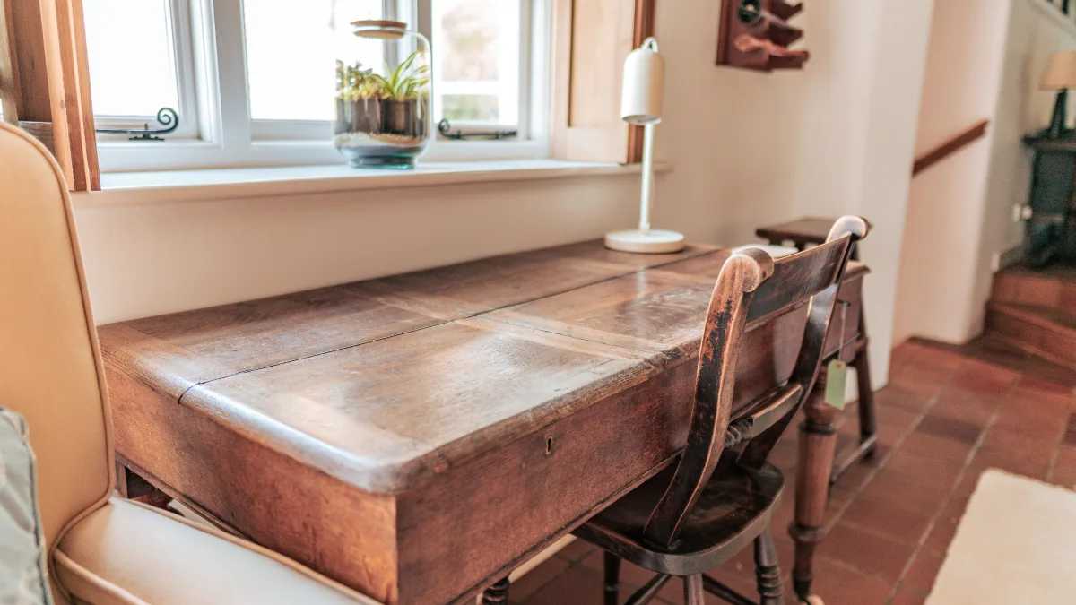 A close view along the warm oak top of the writing desk, the window and a glass terrarium catching the light at the far end