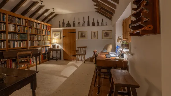The library at Well Cottage in the evening, lamplight warming the bookshelves and a desk lamp pooling light on the writing table