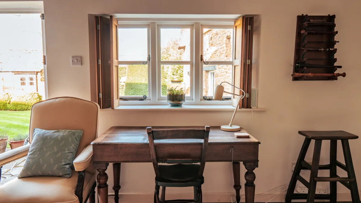 The library writing desk set beneath a deep casement window with the shutters folded back, a reading lamp and an upholstered armchair, looking out over the walled garden