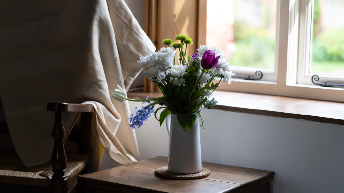 A white jug of garden flowers — white chrysanthemums, a purple tulip and blue hyacinth — on a small oak table by the living-room window