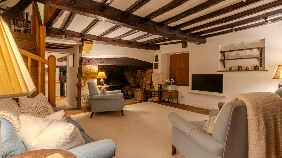 The hearth corner of the living room, a lit lamp glowing on the table beside the inglenook with pale armchairs and a flower jug nearby
