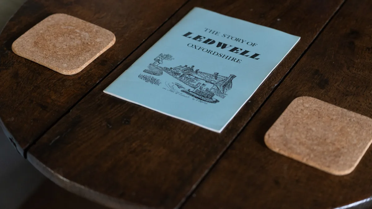 A booklet titled The Story of Ledwell, Oxfordshire, laid between cork coasters on a round oak table