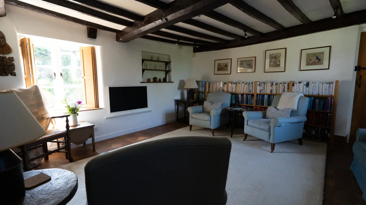 Looking up the lime-washed boxed staircase from the living room, light falling through a deep stone window past an old oak beam to the books on the landing