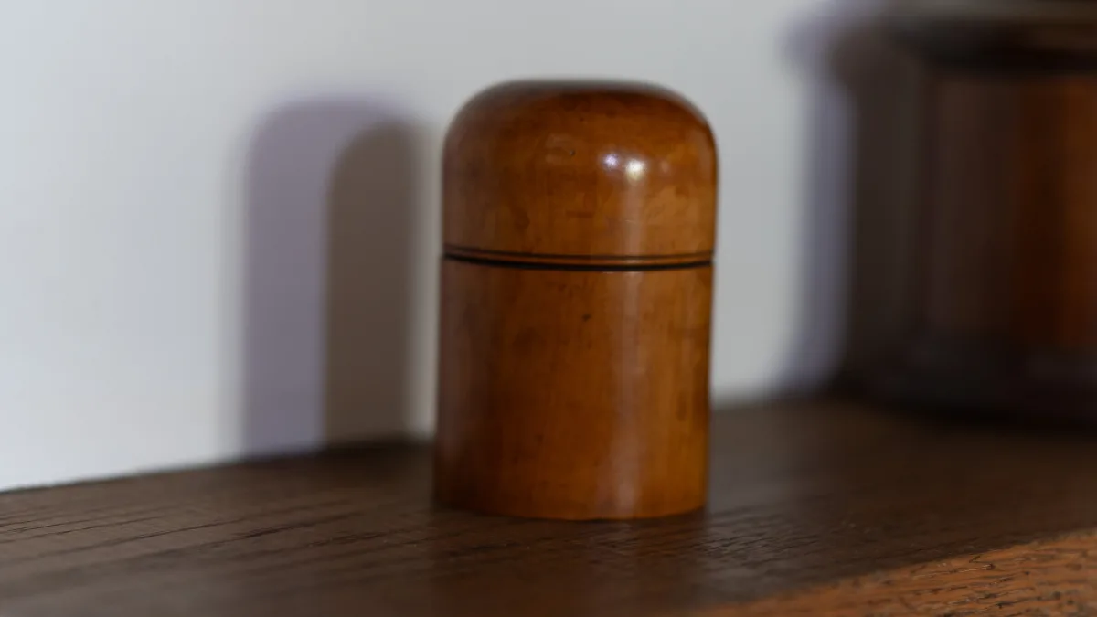 A turned wooden treen canister with a domed lid, resting on a shelf against the white plaster wall