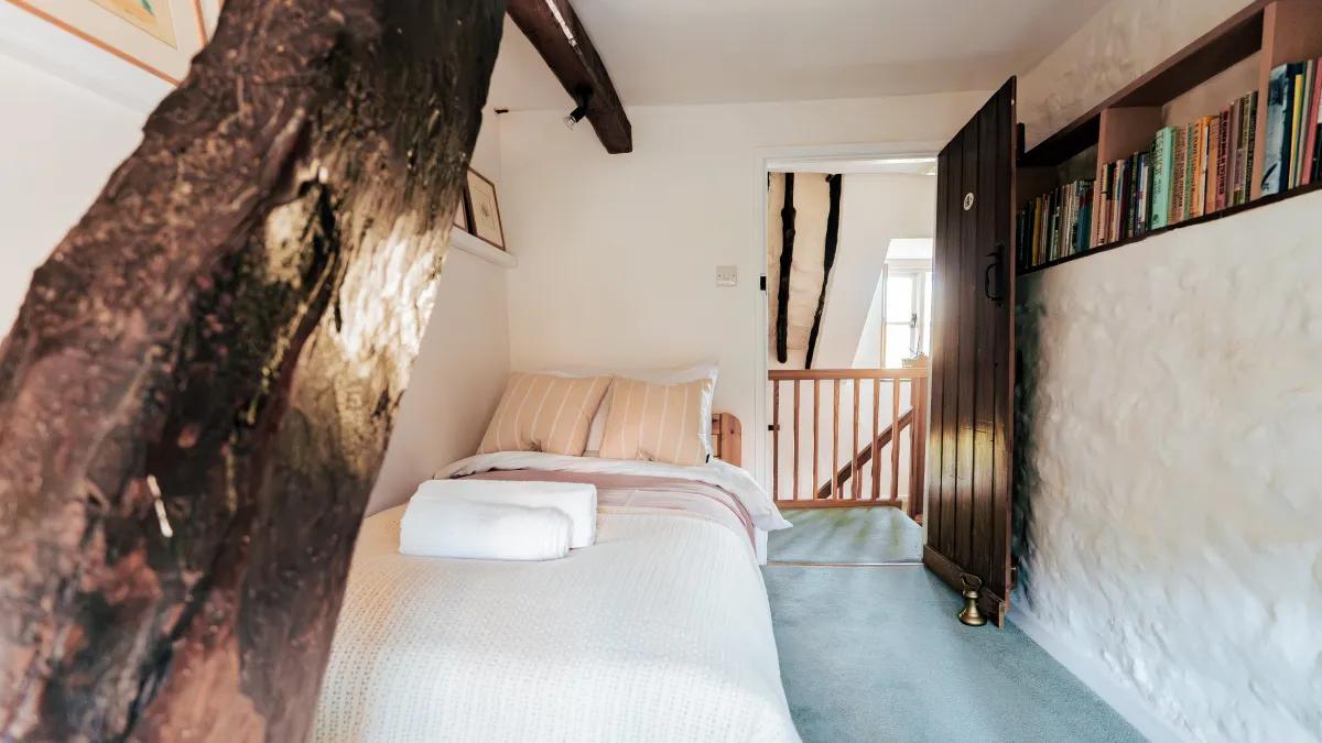 The single bed beneath the great gnarled oak beam that leans across the room, with the bare stone wall and bookshelf beside the open plank door