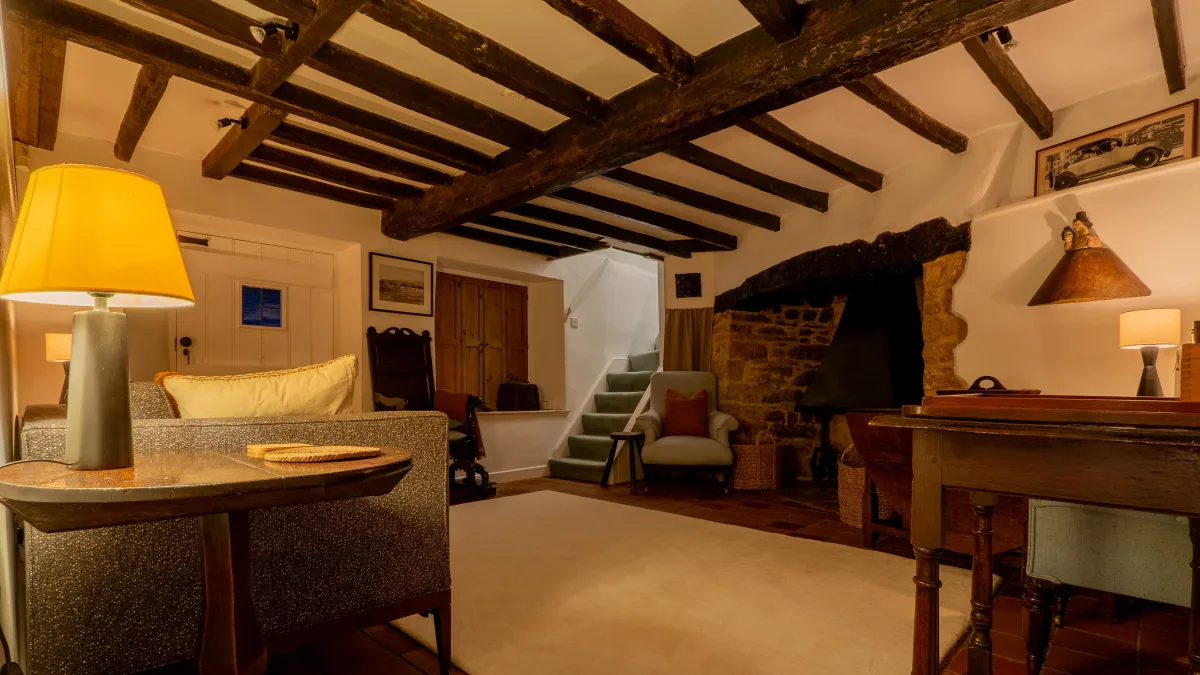 The snug in evening lamplight, table lamps glowing warm over the sofa and gateleg tables, the inglenook and winding stair in shadow beneath the dark beams