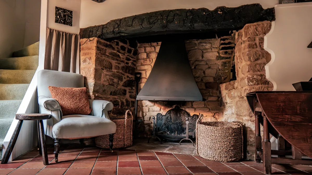 The snug's deep stone inglenook with its heavy timber lintel and iron hood, a soft armchair drawn up beside it and woven log baskets on the quarry-tiled floor