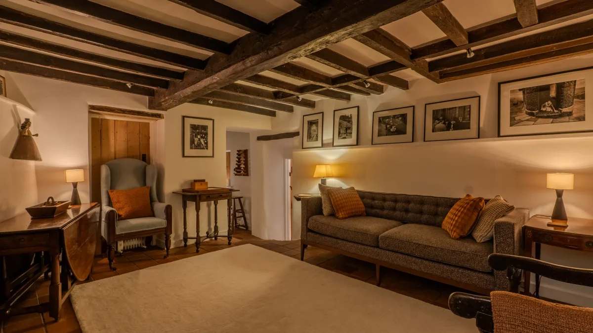 The tweed sofa in the snug dressed with burnt-orange cushions beneath a row of framed black-and-white prints, a wing chair and gateleg tables with table lamps to either side