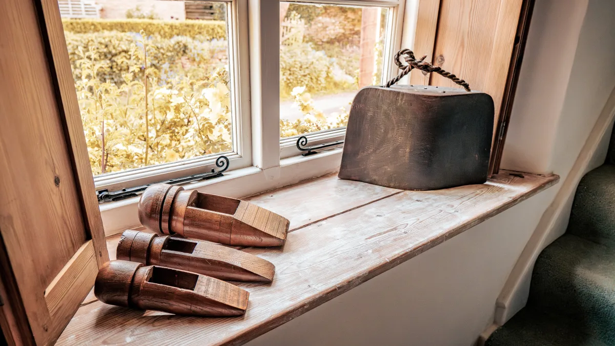 A close view along the worn pine windowsill, the wooden shoe lasts and rope-handled cow bell lit by soft daylight from the garden beyond