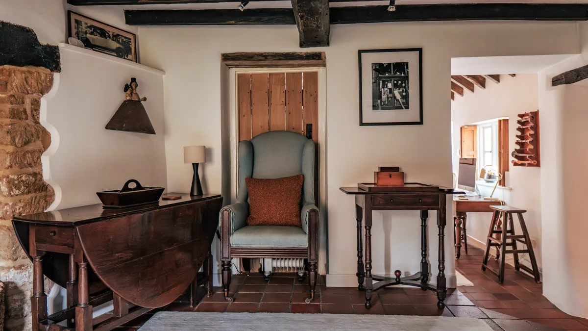 A pale wing chair set in a panelled-door alcove beside a drop-leaf table and the bare stone wall, with a glimpse through to the study beyond