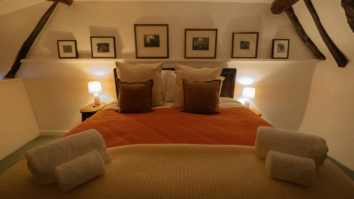 The bed seen square from the foot, twin bedside lamps lit beneath the row of framed prints, rolled white towels set out on the orange and gold blankets