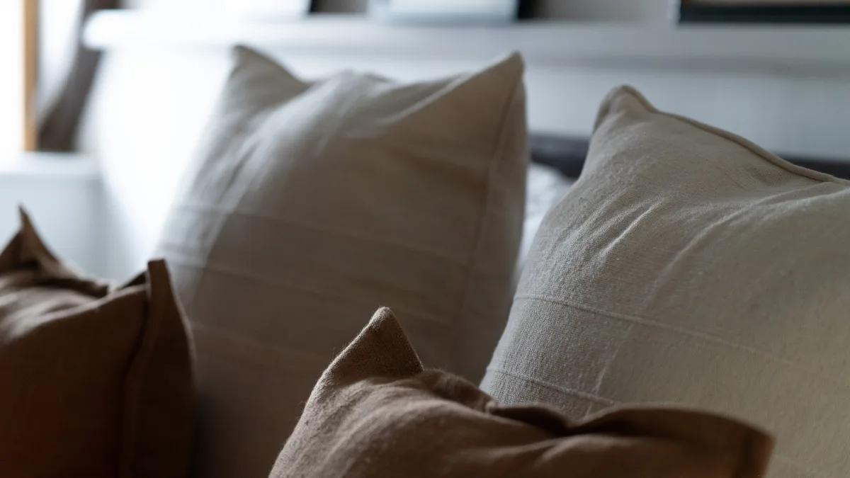 A soft-focus row of oatmeal and tan linen cushions on the super king bed, the shuttered window glowing pale behind
