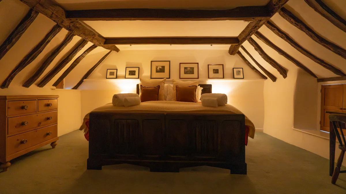 The pine dresser with its turned knobs standing beside the carved foot of the super king bed, beams sweeping down to the corner of the lamplit room