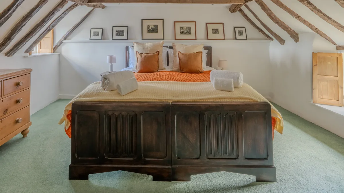 The carved panelled footboard of the super king bed in daylight, the pine dresser to one side and the shuttered window glowing in the bright corner beyond